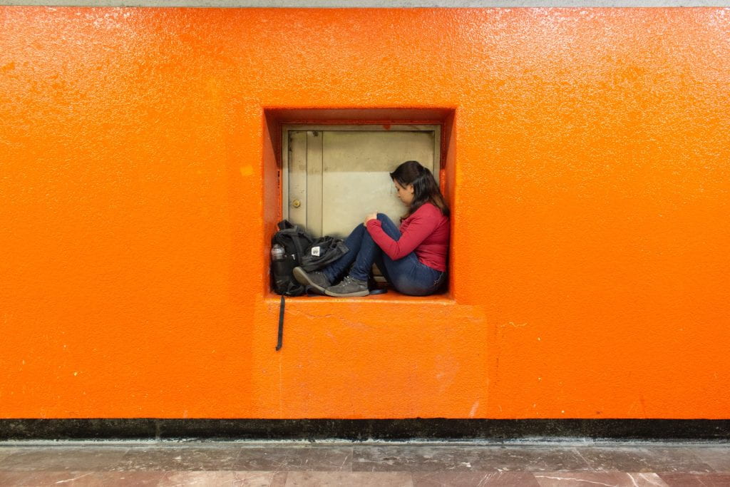 Artwork: Orange wall with a cut out cube and a woman inside with a backpack.